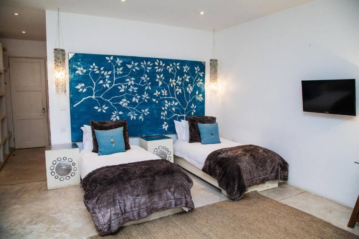 A bedroom with two beds, a large blue wall art, a flat-screen TV, and a modern light fixture.