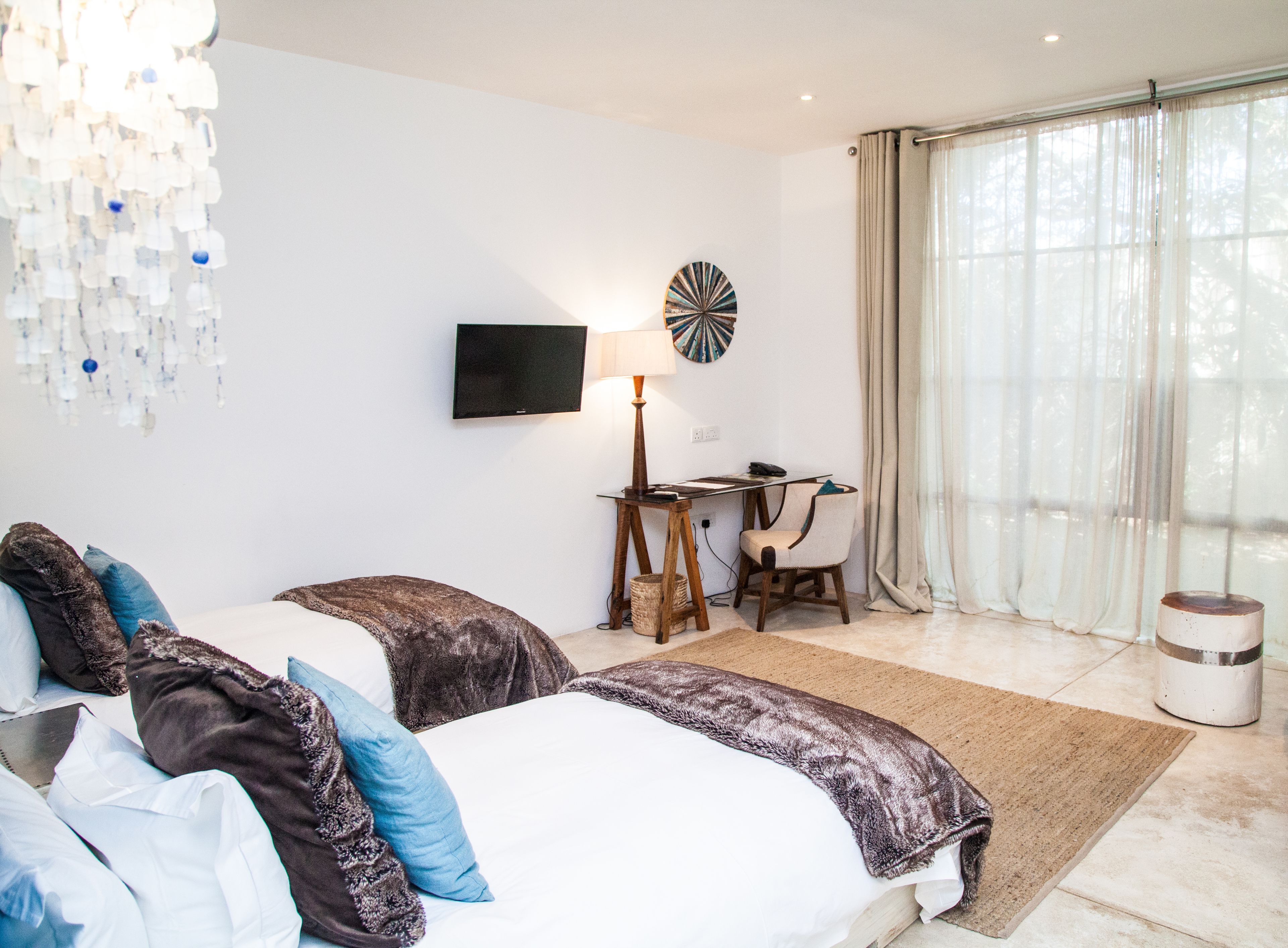 A hotel room with two beds, a desk with a lamp, a TV, and a large window with curtains.