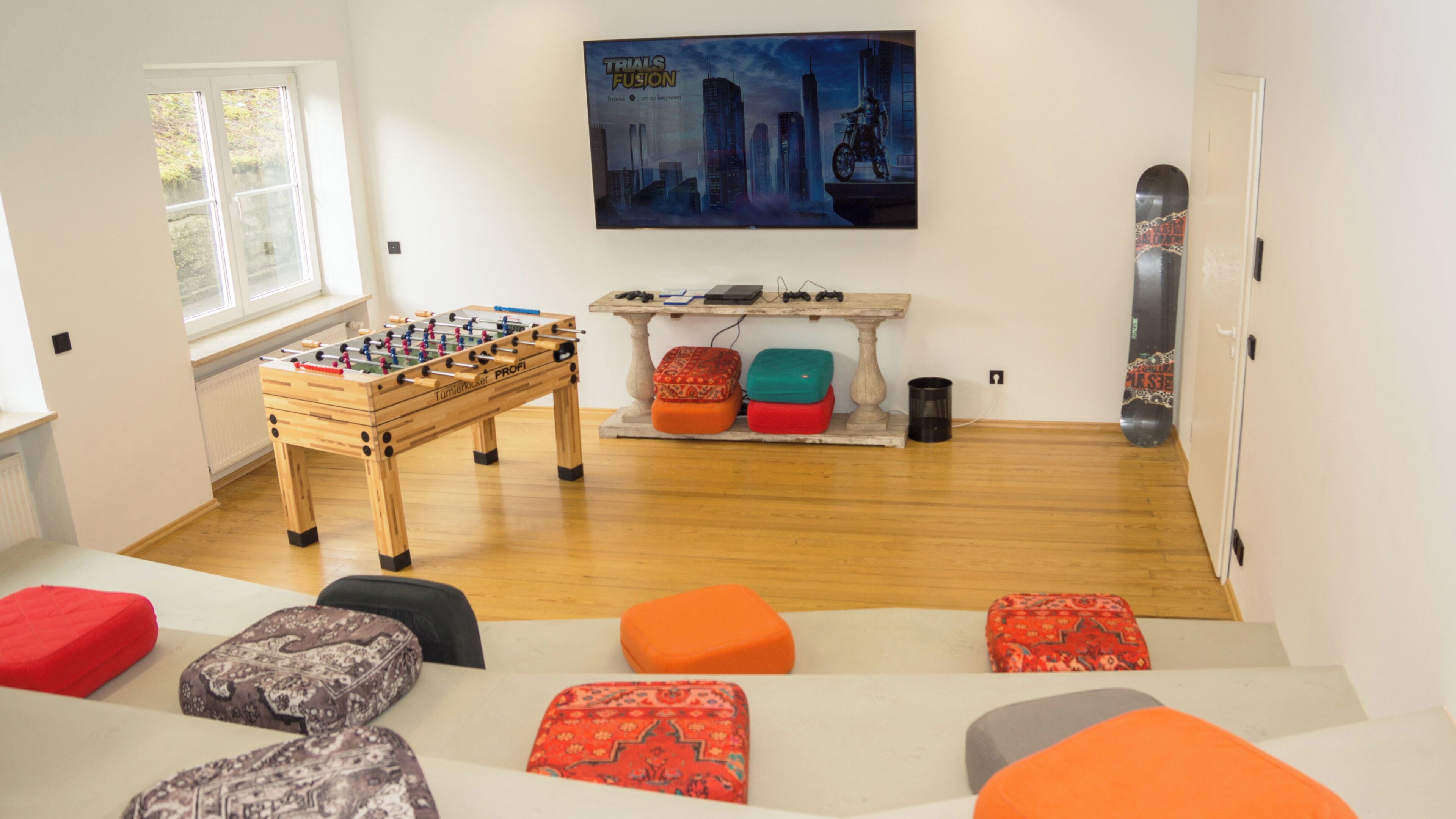A room with a foosball table, colorful ottomans, a mounted TV, and a door with a skateboard.