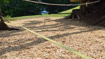 Un parque infantil con un columpio de cuerda, suelo de mantillo y un árbol al fondo.