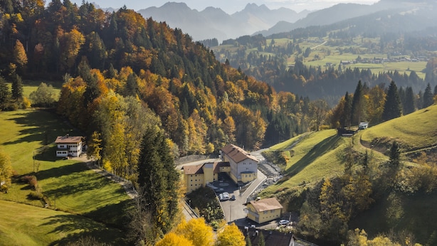 A large building with multiple sections, surrounded by lush green fields and trees with autumn foliage.