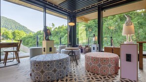 A modern interior with round ottomans, a wooden dining table, and chairs. Large windows offer a view of a forested hillside.