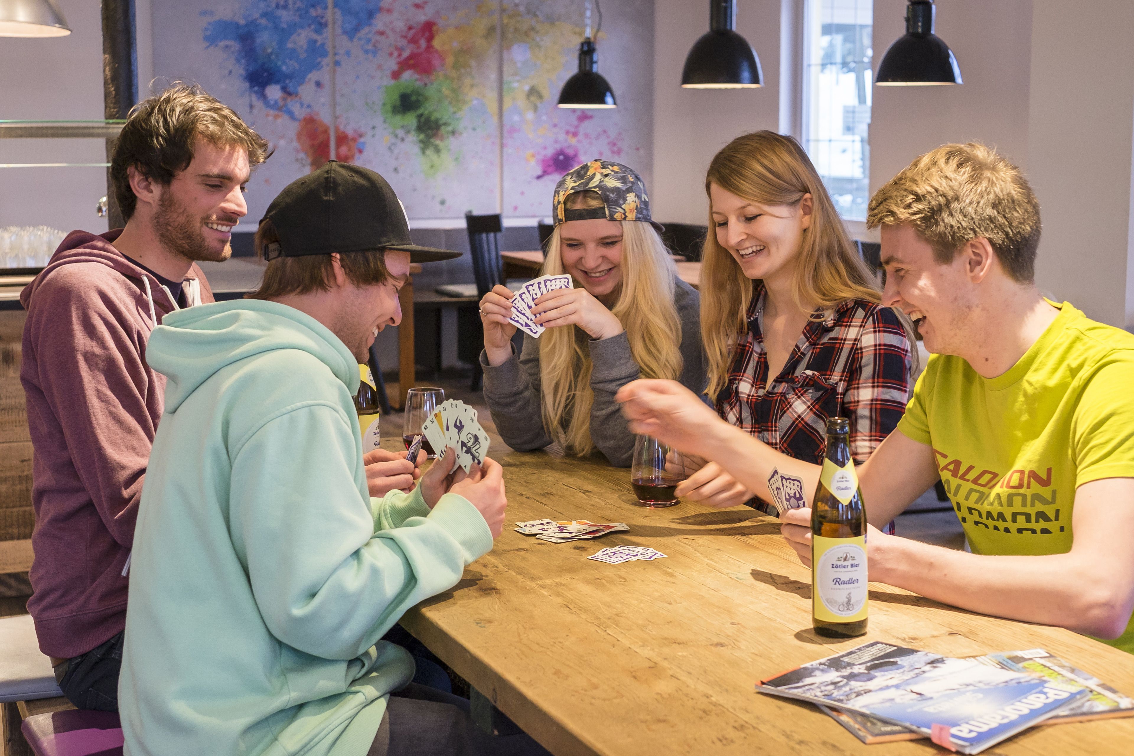 A group of people playing cards at a wooden table with a bottle of beer and playing cards on it.