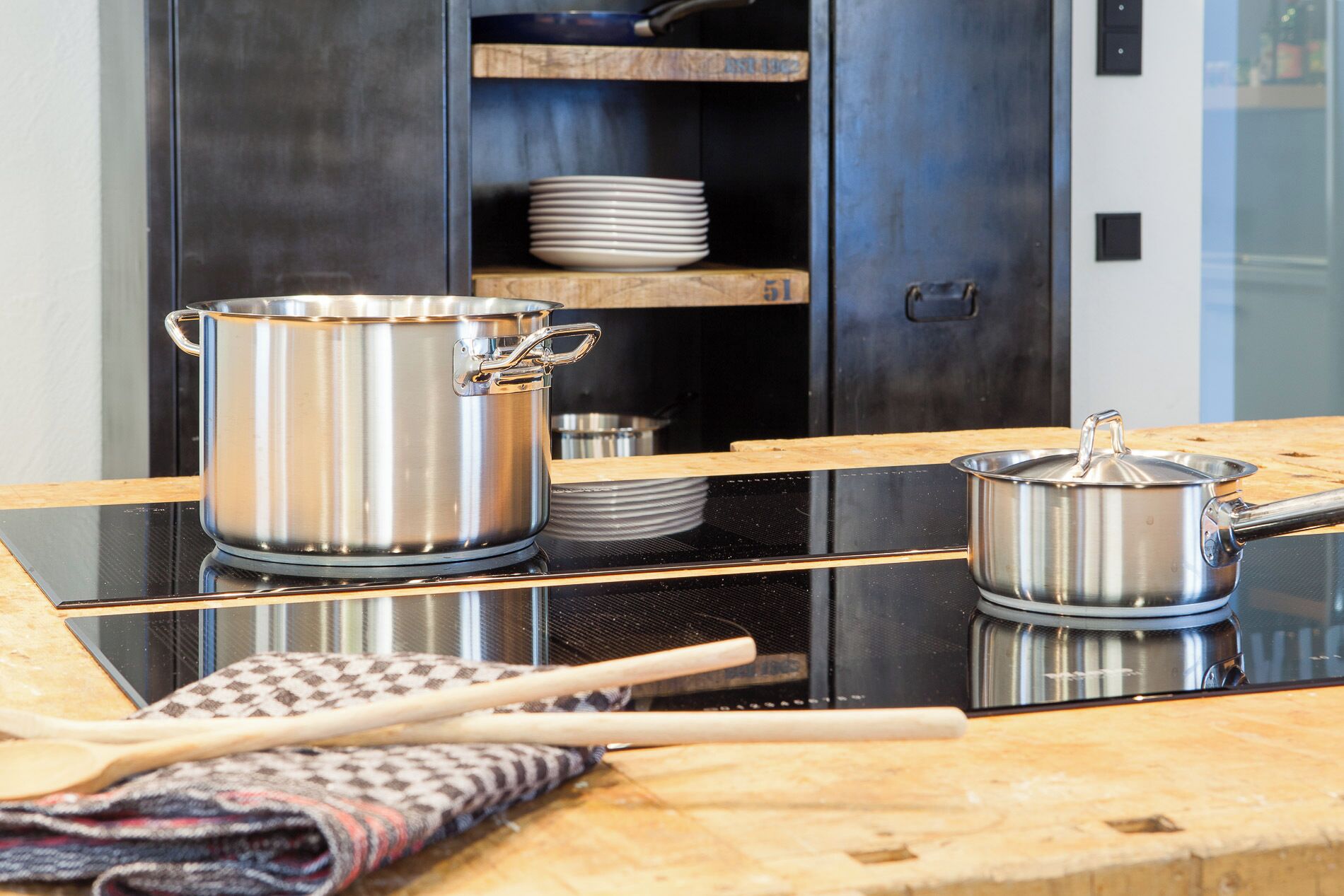 Una cocina con ollas de acero inoxidable sobre una encimera negra, un textil tejido y un estante de madera con platos.