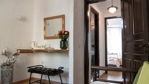 Een kamer met een bed, een klein tafeltje met een kopje en een krant, een spiegel, een vaas met bloemen, en een deur die leidt naar een andere kamer.