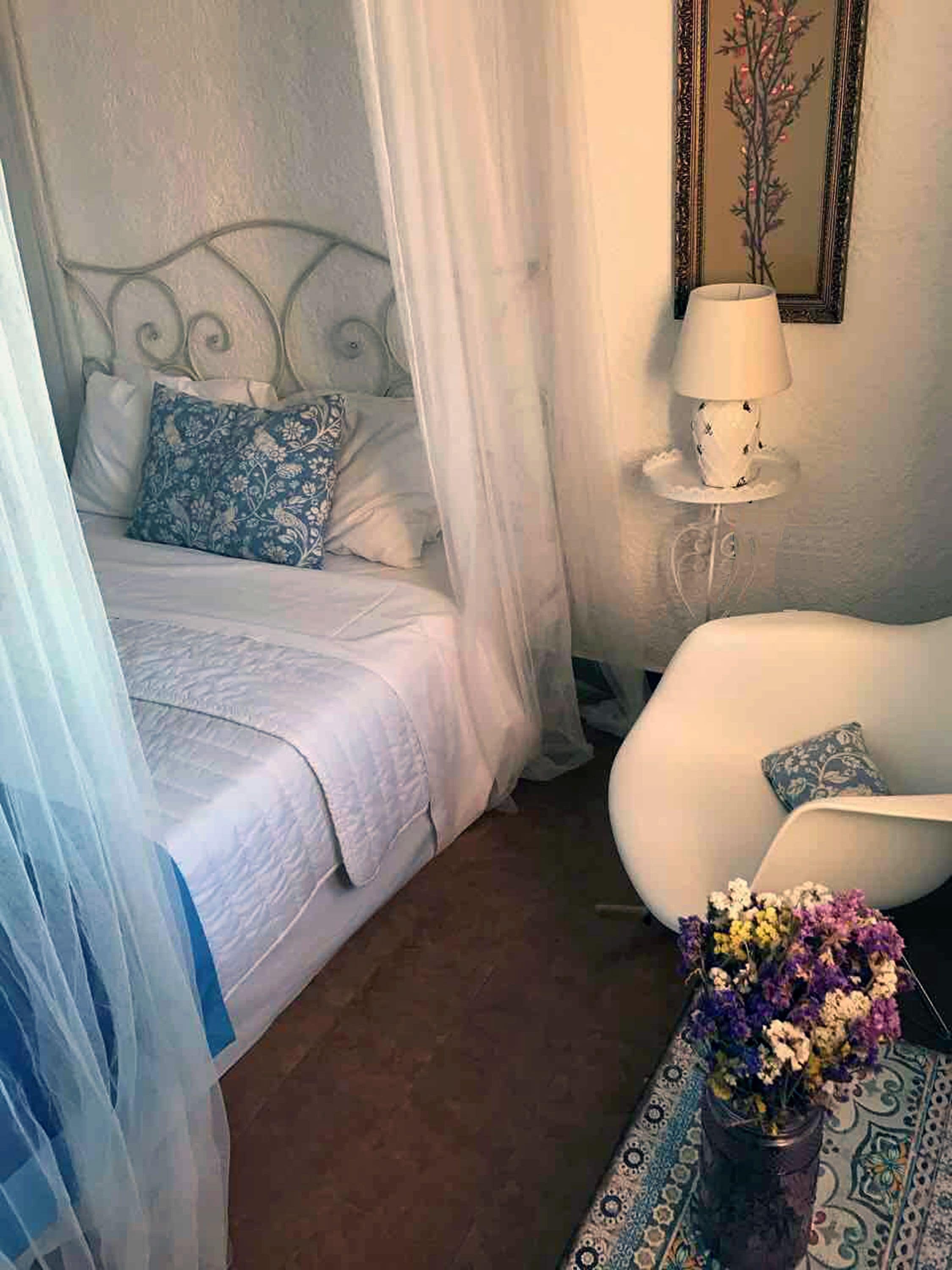 A bedroom with a bed, a bedside table with a lamp, a chair, and a vase of flowers.
