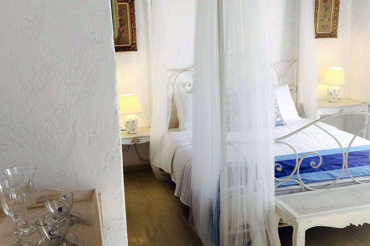 Ein Schlafzimmer mit Himmelbett, weißen Wänden und Holzboden. Es gibt zwei Nachttische mit Lampen sowie ein kleines Regal mit Gläsern.