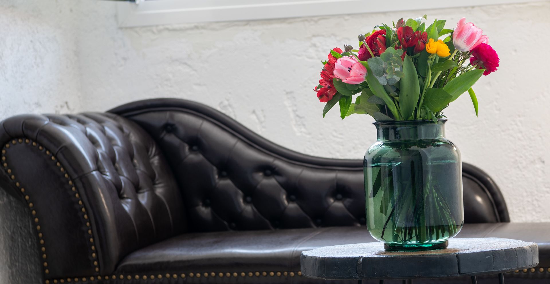 Een donkerbruine leren bank met een gestuikte rugleuning en gouden studs, geplaatst tegen een witte muur. Voor de bank staat een groene glazen vaas met een bloemenboeket op een klein, rond tafeltje.