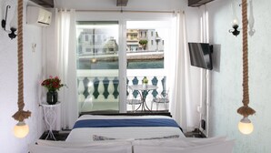 Hotelkamer met een bed, een tafeltje met stoelen, een televisie en een balkon met uitzicht op het water en gebouwen.