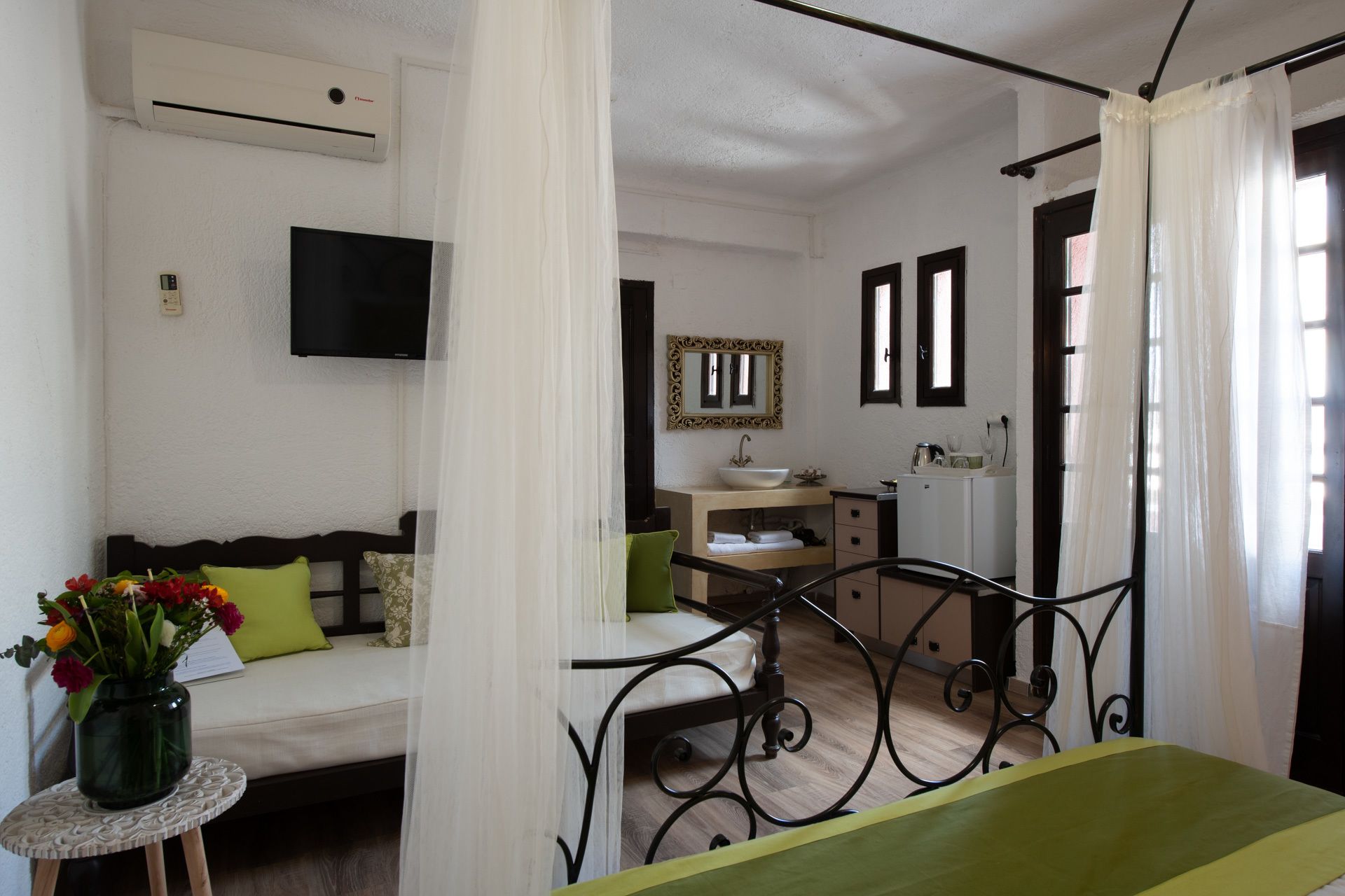 A room with a bed, a small table with a vase of flowers, a desk with a computer, a bathroom with a sink and mirror, and a TV mounted on the wall.