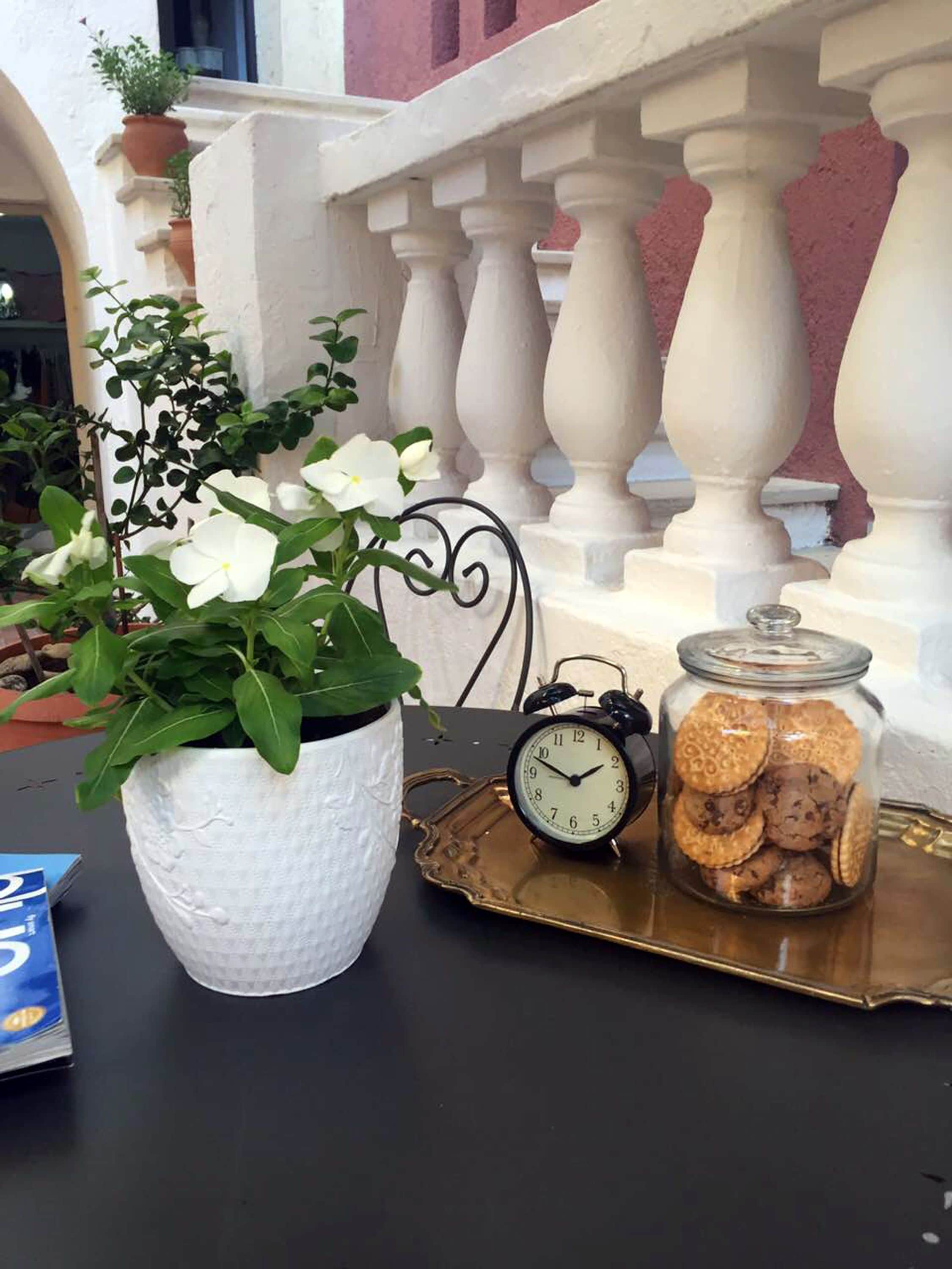 A table with a white textured pot containing white flowers, a black alarm clock, and a glass jar filled with cookies on a gold tray.