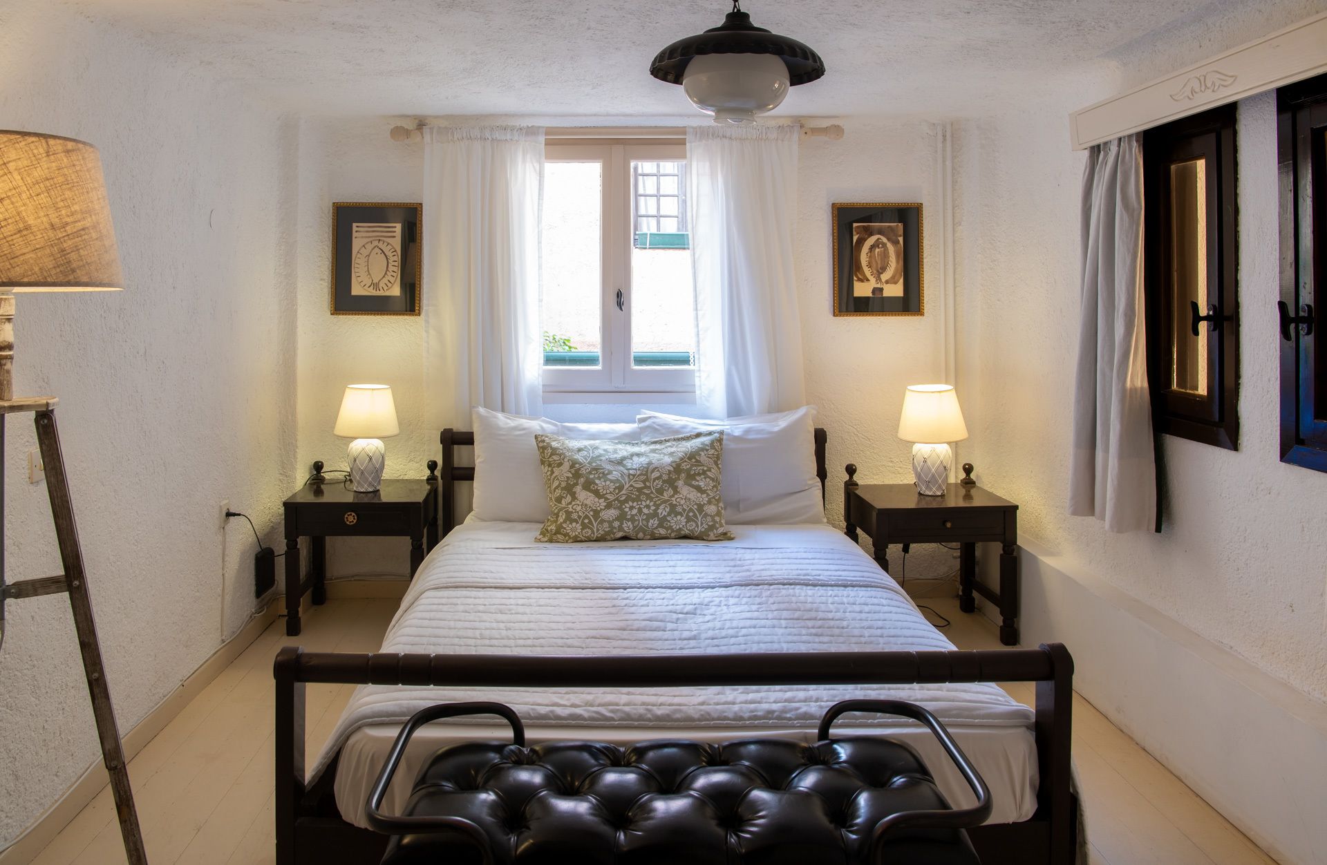A bedroom with a bed, two bedside tables with lamps, a window with curtains, and a wall-mounted picture frame.