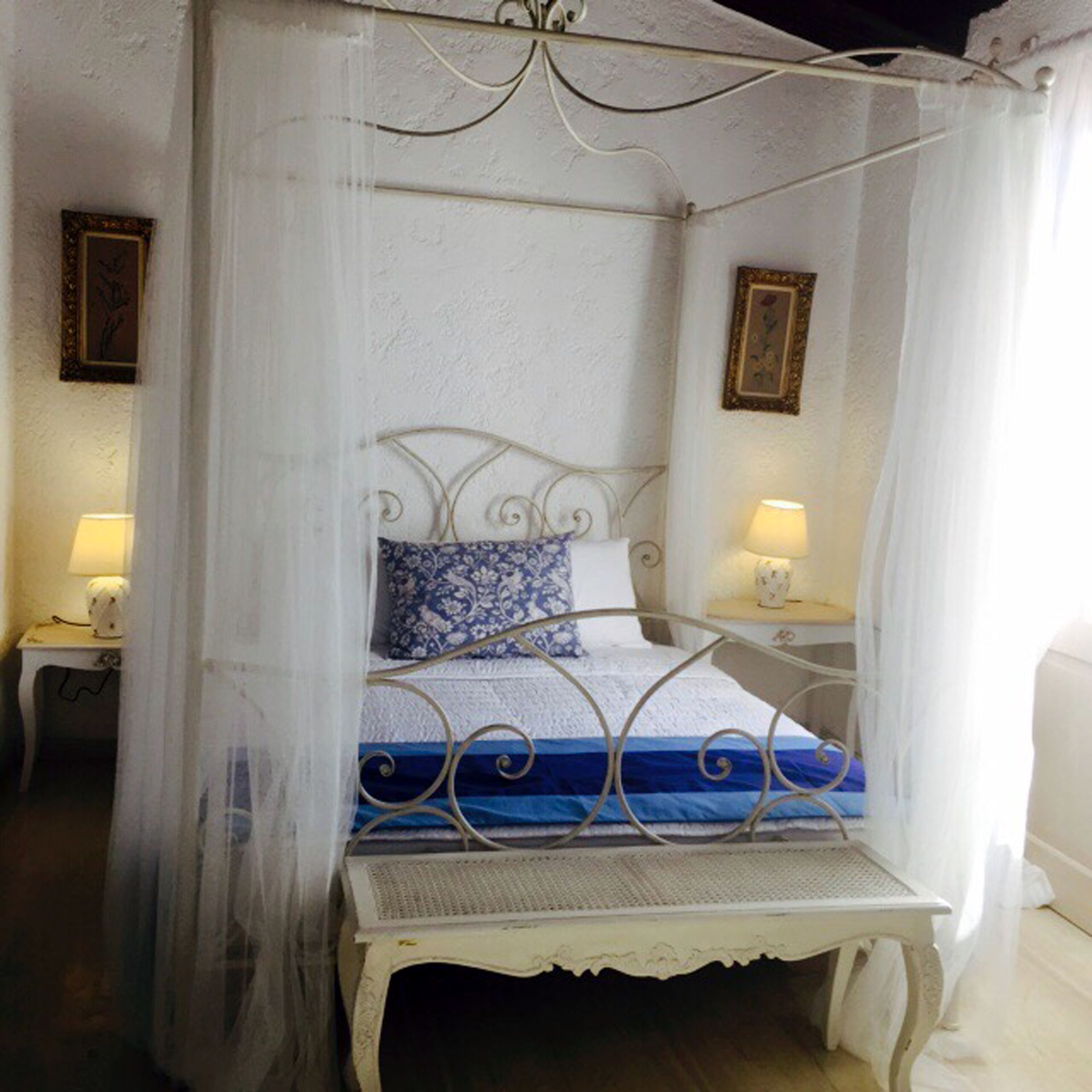 A four-poster canopy bed with a white metal frame, a blue and white patterned pillow, and a white bedspread. There are two bedside tables with lamps on either side of the bed.