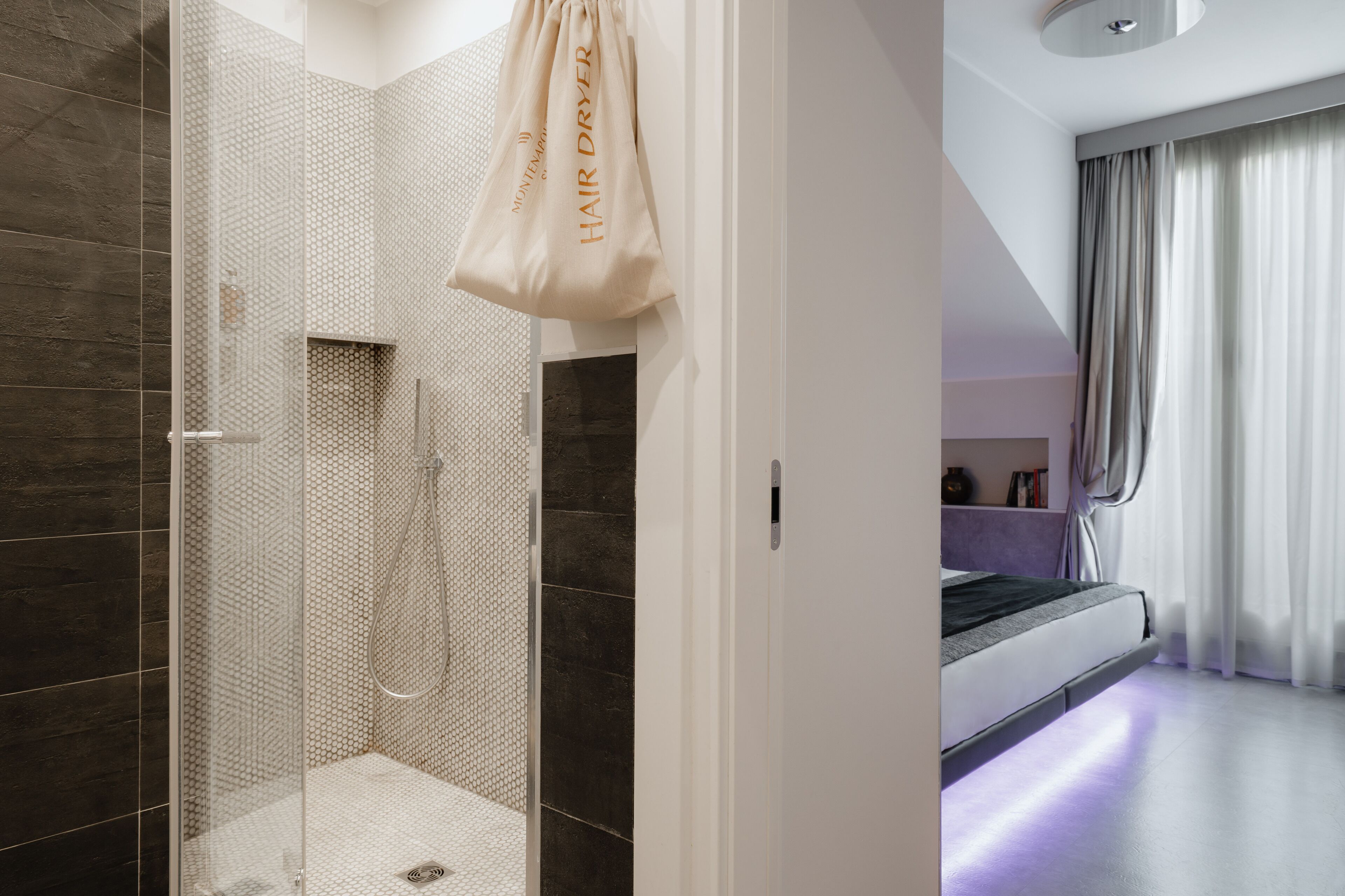 Um banheiro moderno com box de vidro, uma bolsa bege para secador de cabelo e um quarto com cama e cortinas.