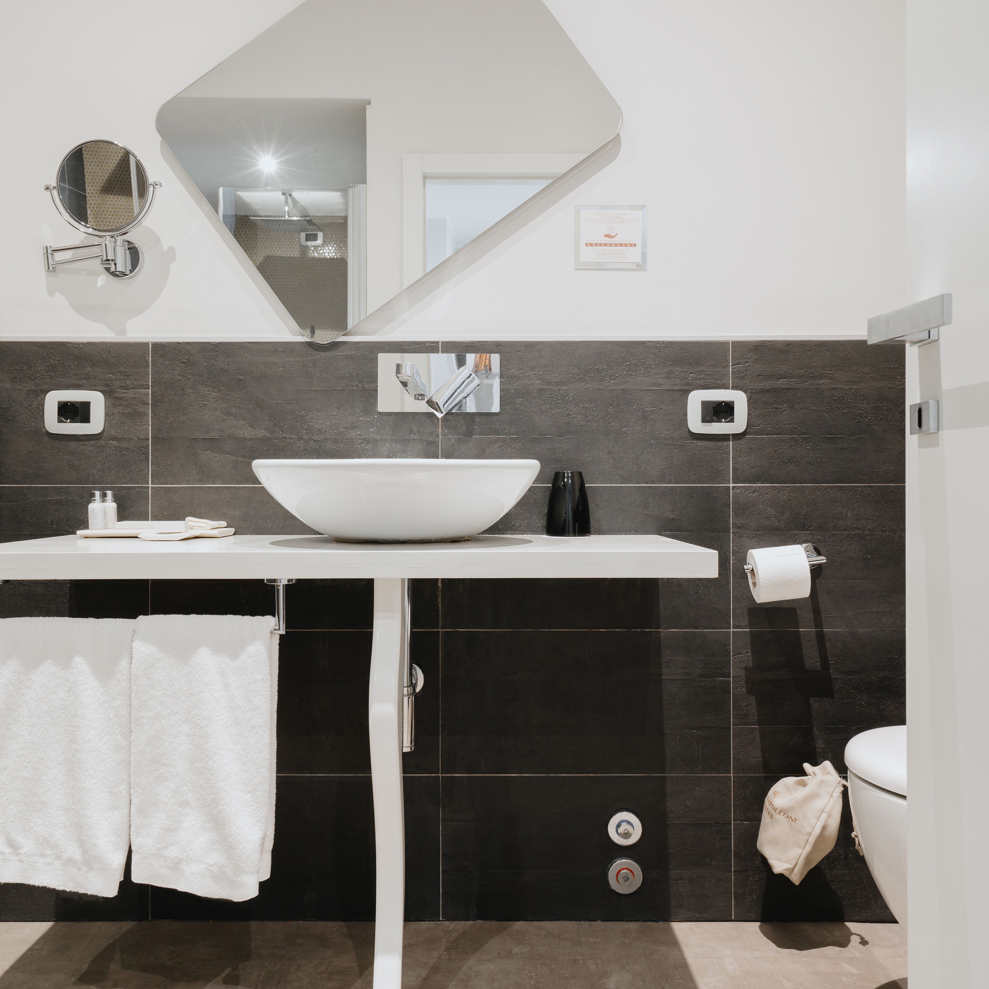 A modern bathroom with a white sink, a round mirror, and a large mirror above the sink. There are two towel racks with white towels, a toilet, and a paper towel dispenser.