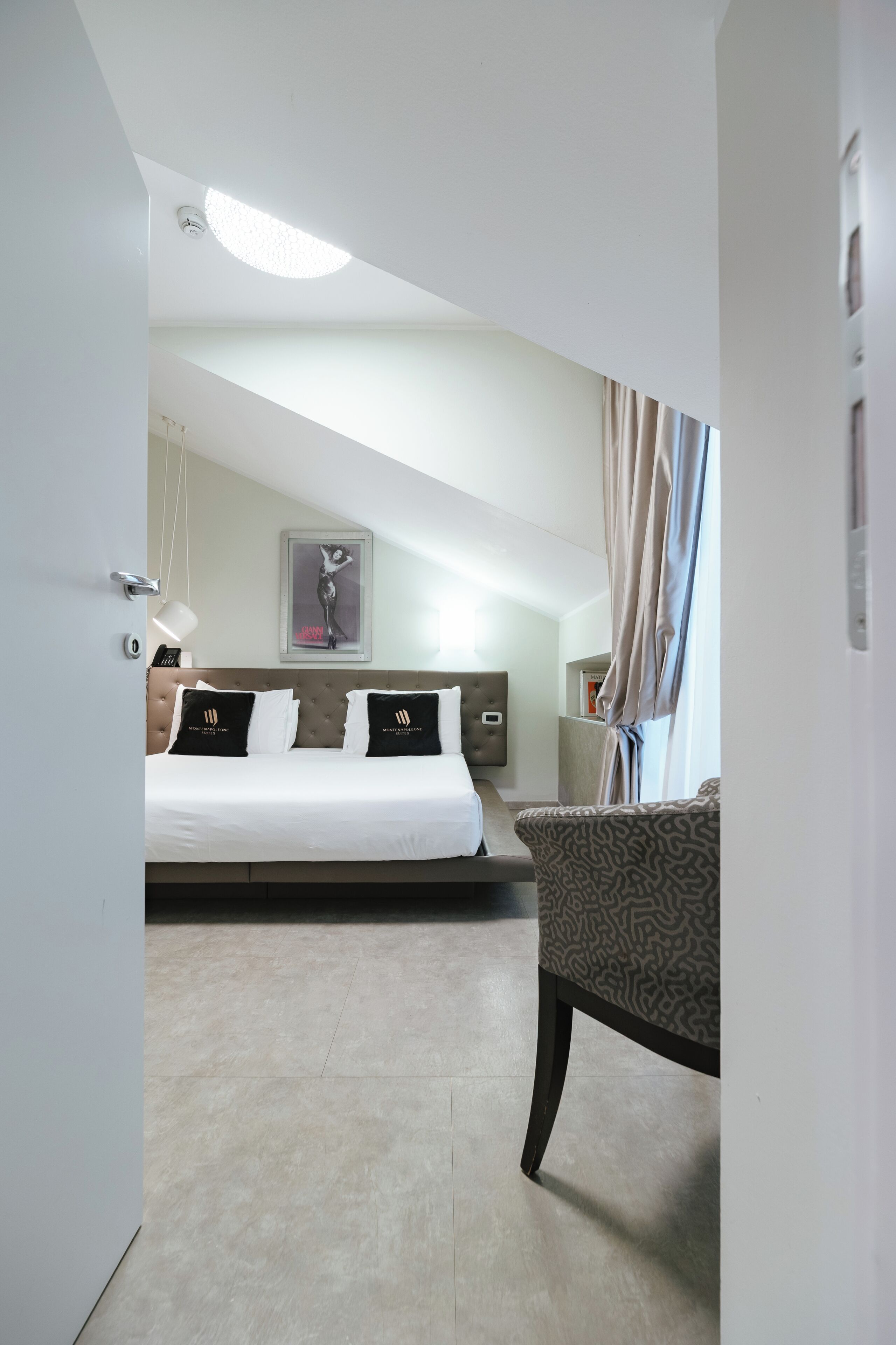 A hotel room with a bed, a chair, a ceiling light, a wall-mounted lamp, and a framed picture.