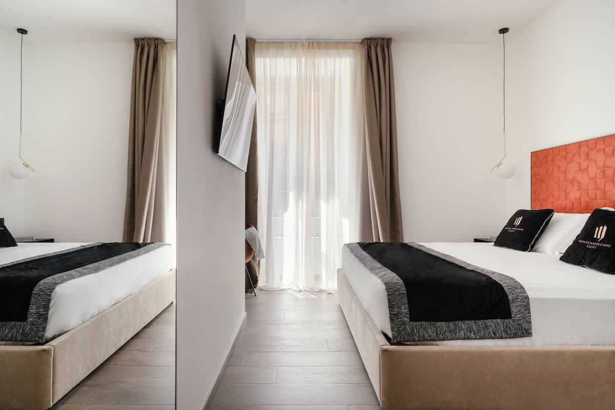 Habitación de hotel con dos camas, un ventanal con cortinas translúcidas, un espejo y una pared de acento en color rojo.