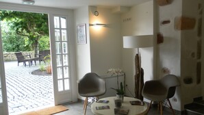 A modern living room with a round table, two chairs, a lamp, and a door leading to an outdoor patio with a brick floor and outdoor furniture. - Hôtel-Résidence Alaia (Ascain)