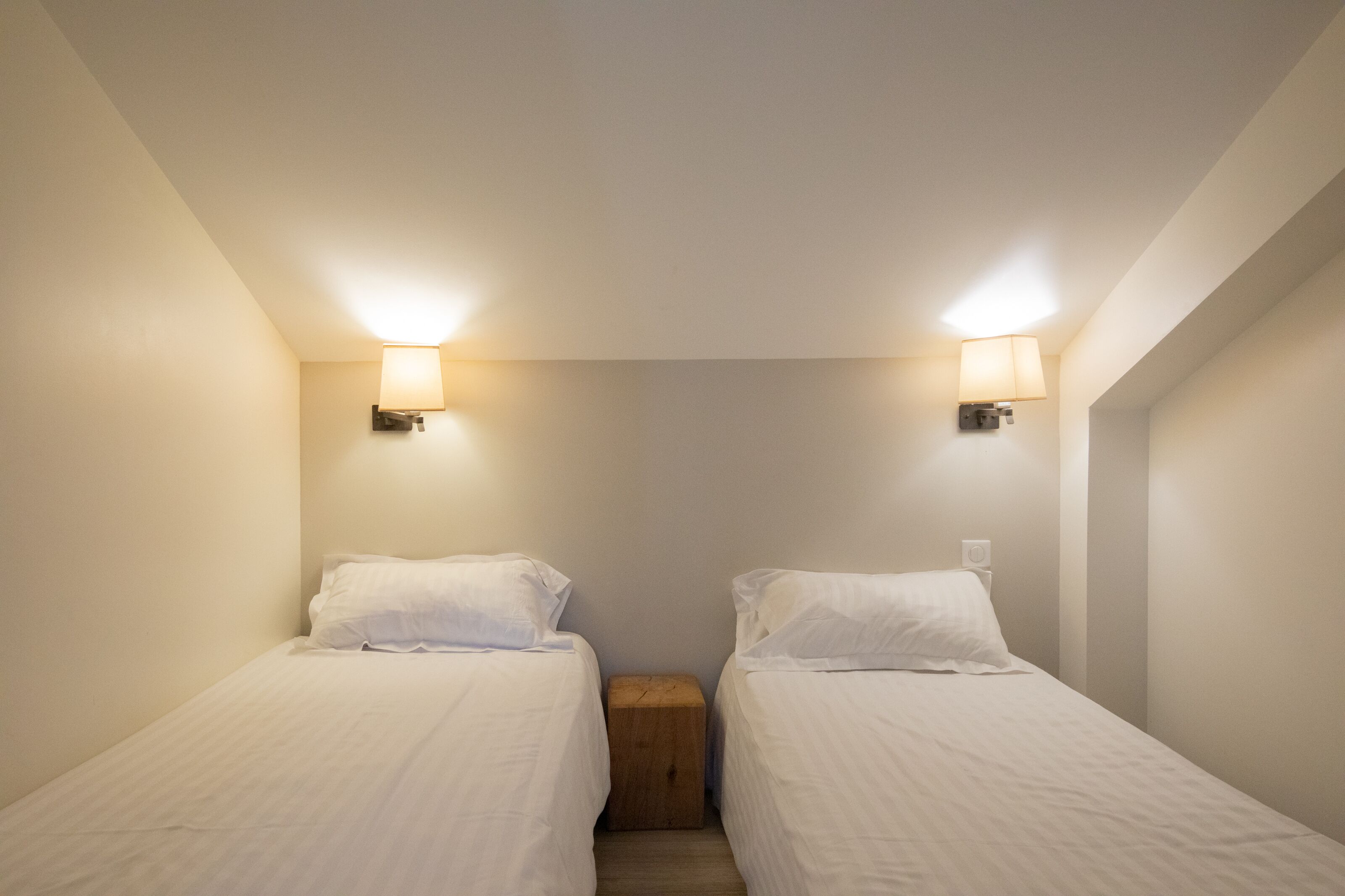A small, well-lit bedroom with two single beds, a wooden nightstand, and wall-mounted lamps.