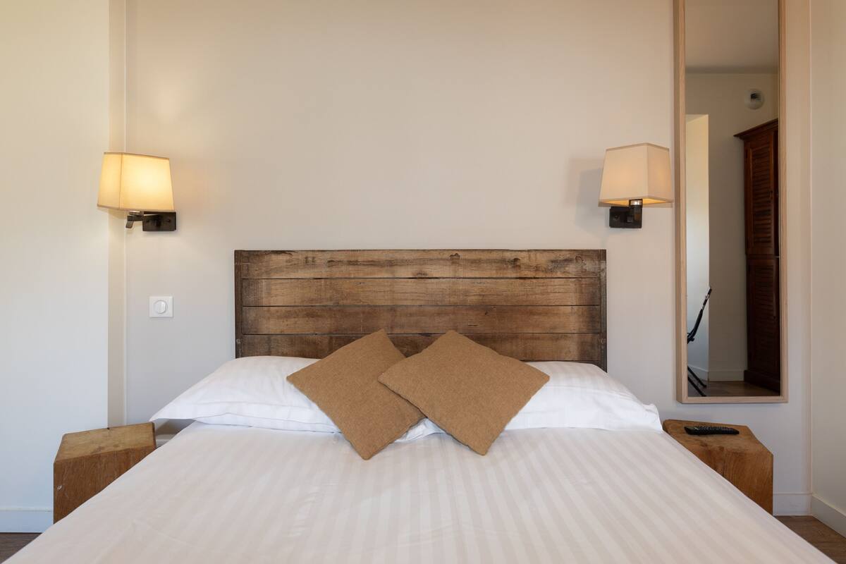 A neatly made bed with two pillows, a wooden headboard, bedside lamps, and a nightstand with a remote control.