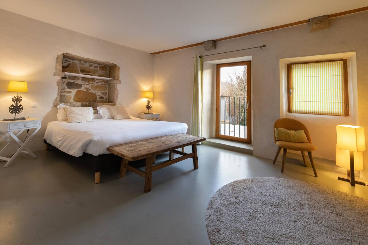 A bedroom with a stone wall, a bed, a bench, a chair, and two bedside lamps.