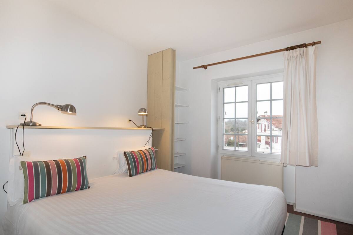 A bedroom with a bed, two bedside lamps, a window with curtains, and a view of buildings outside.