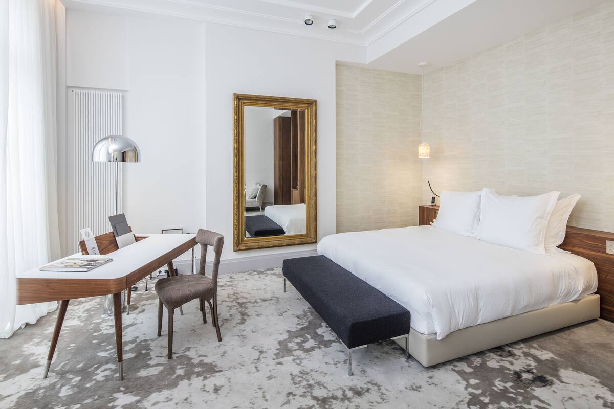 Una habitación de hotel con cama, mesita de noche, silla, escritorio y un espejo grande.