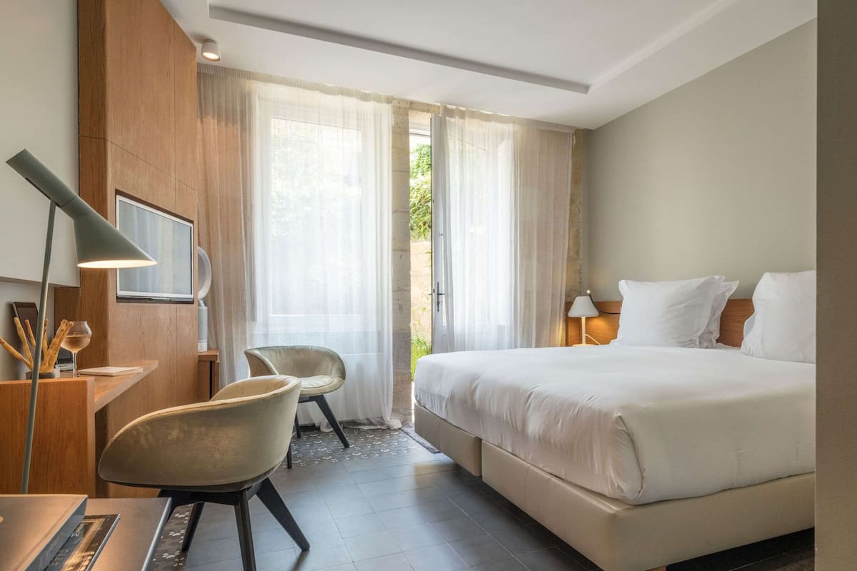 Habitación de hotel con cama, escritorio con lámpara, una silla y un ventanal con cortinas translúcidas.