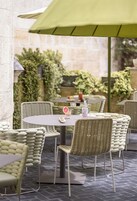 An outdoor patio with wicker chairs and tables, a green umbrella, and a stone wall in the background.