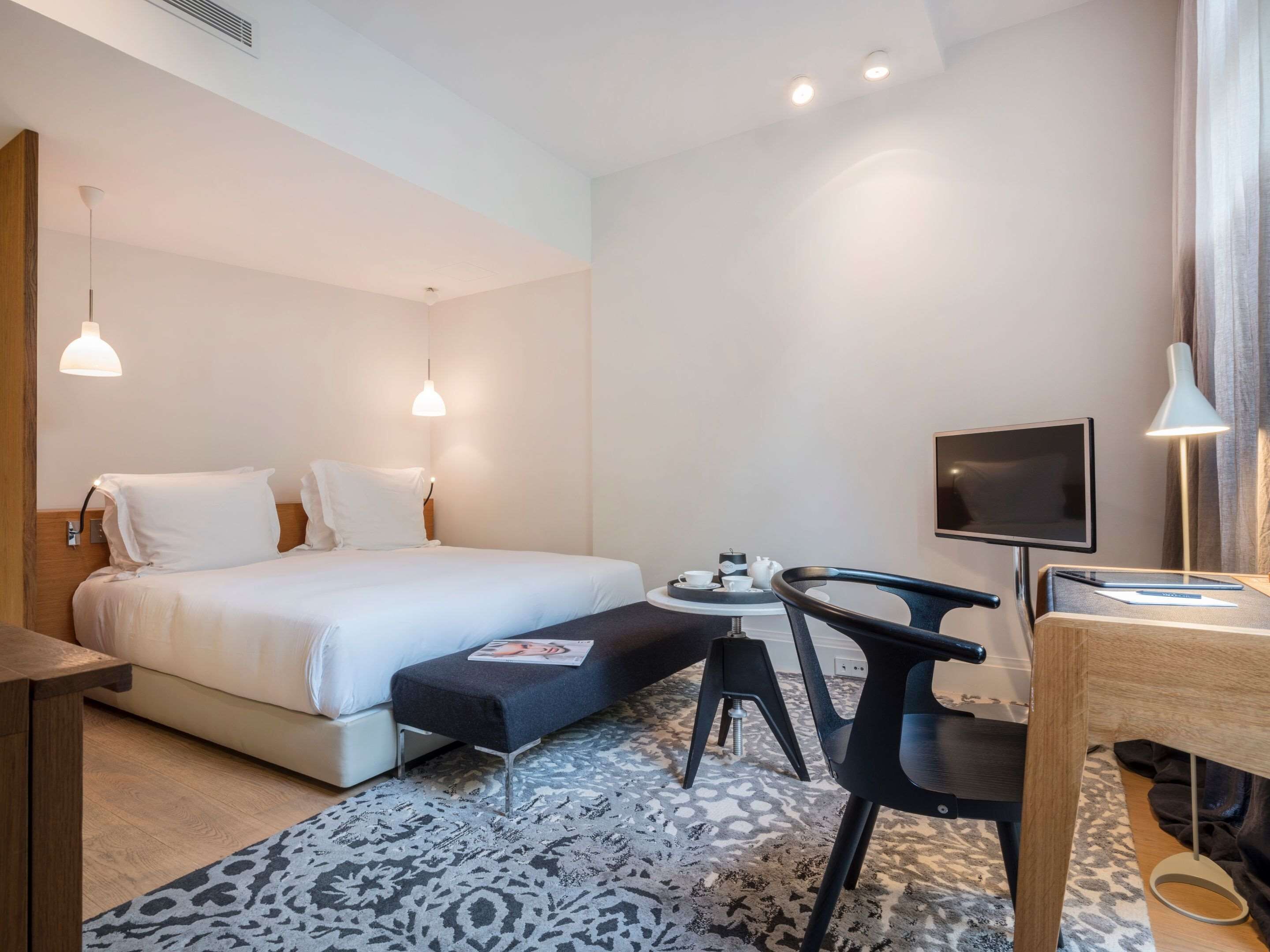A hotel room with a bed, bedside table, chair, desk, TV, and a small table with cups and a teapot.