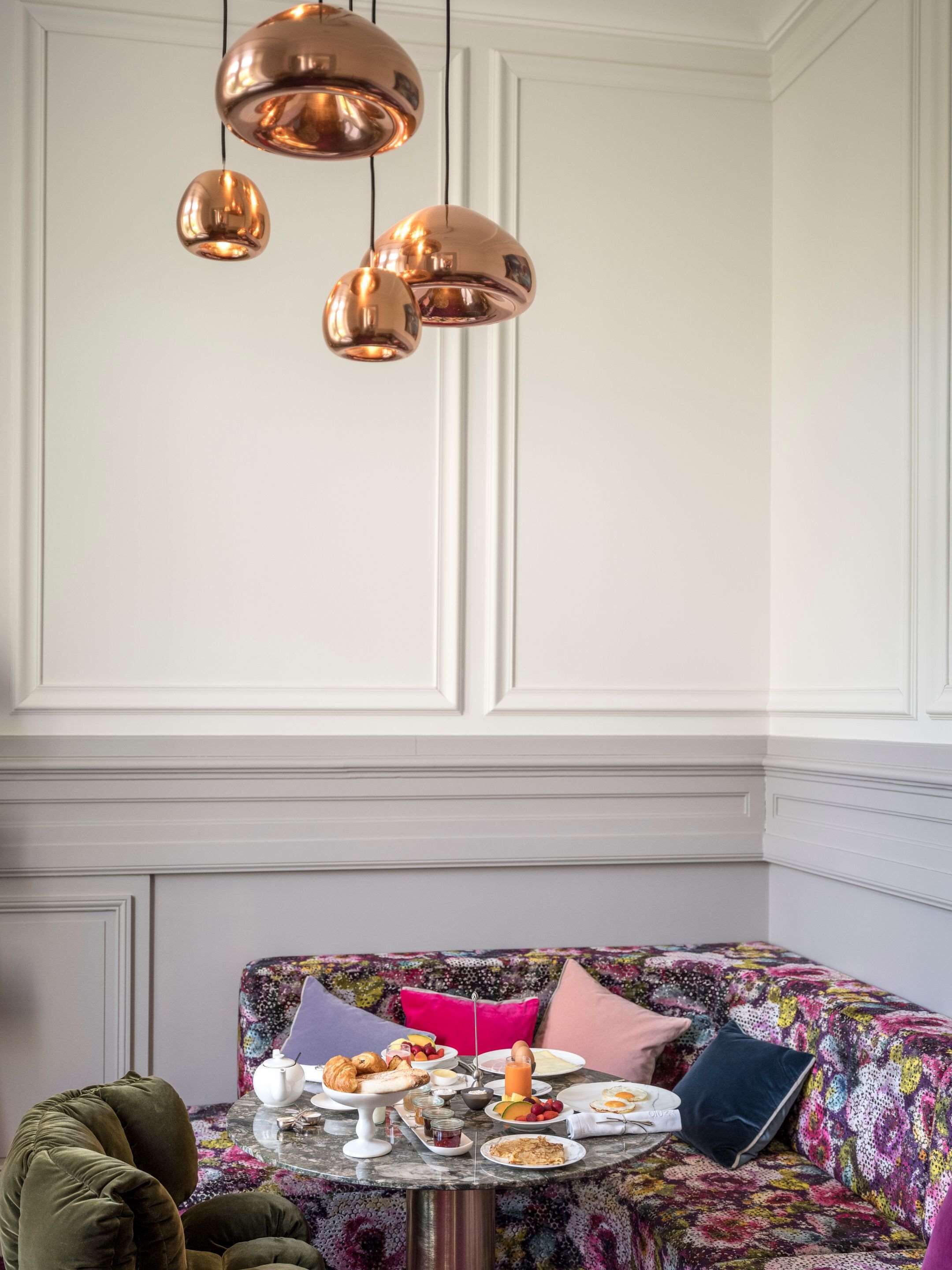 Un comedor con una mesa redonda dispuesta para el desayuno, un sofá estampado y una lámpara colgante decorativa de cobre.
