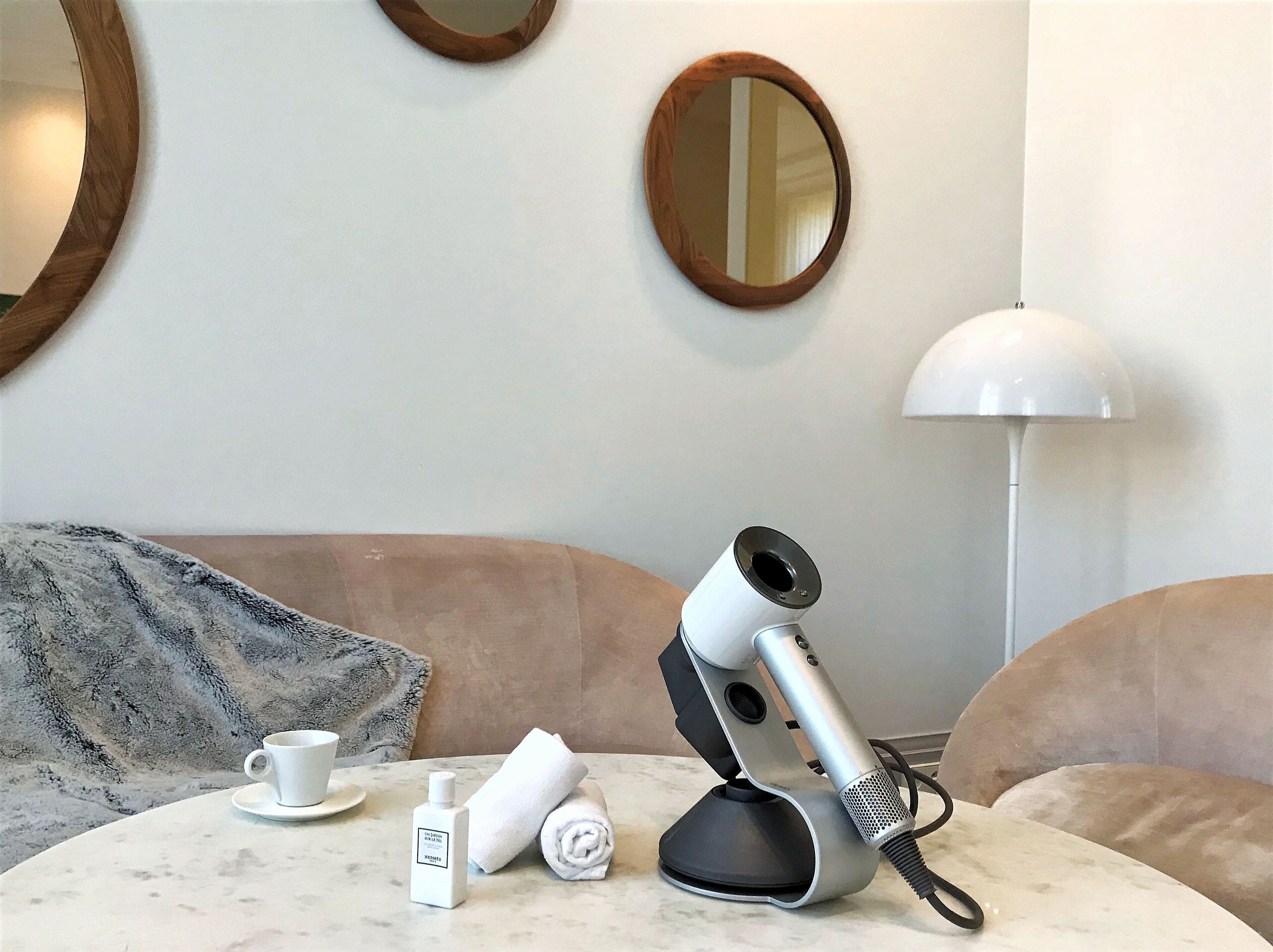 A room with a round mirror, a white lamp, a hairdryer, a cup, and a bottle on a table.