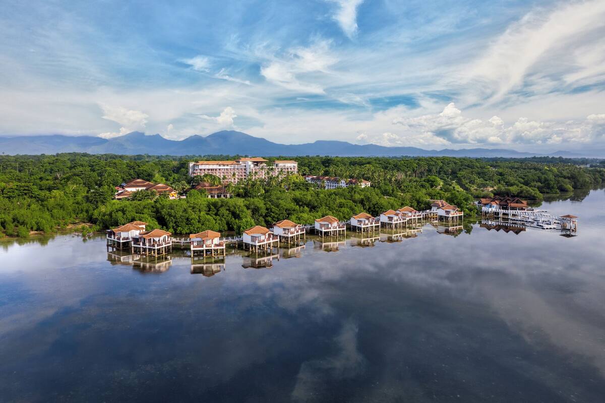 Een pand aan het water met meerdere bungalows op palen, een centraal gebouw en weelderige begroeiing.