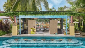 Un bar situé au bord de la piscine, dans un cadre tropical, avec une piscine ornée de motifs en mosaïque et un espace salon couvert agrémenté de plantes grimpantes.