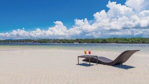 Une plage avec un fauteuil de plage et une table sur laquelle sont posées deux boissons, offrant une vue sur une mer calme et une côte lointaine parsemée de bâtiments.