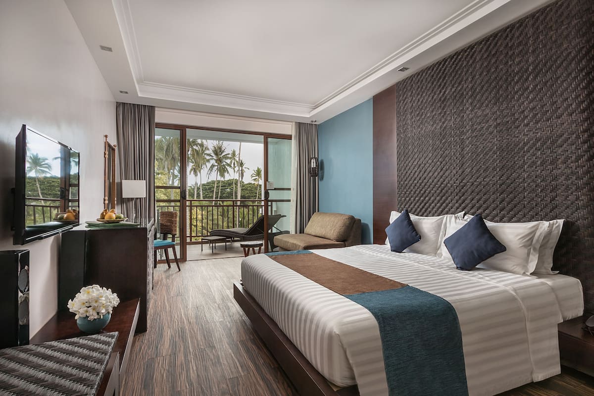 Een hotelkamer met een bed, een televisie, een bureau, een stoel, een balkon met uitzicht op palmbomen en een geweven hoofdbord.