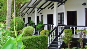 A building with a covered porch, white walls, and black trim. There are steps leading up to the porch, and the area is surrounded by greenery. - Hotel Flower Garden (Unawatuna)