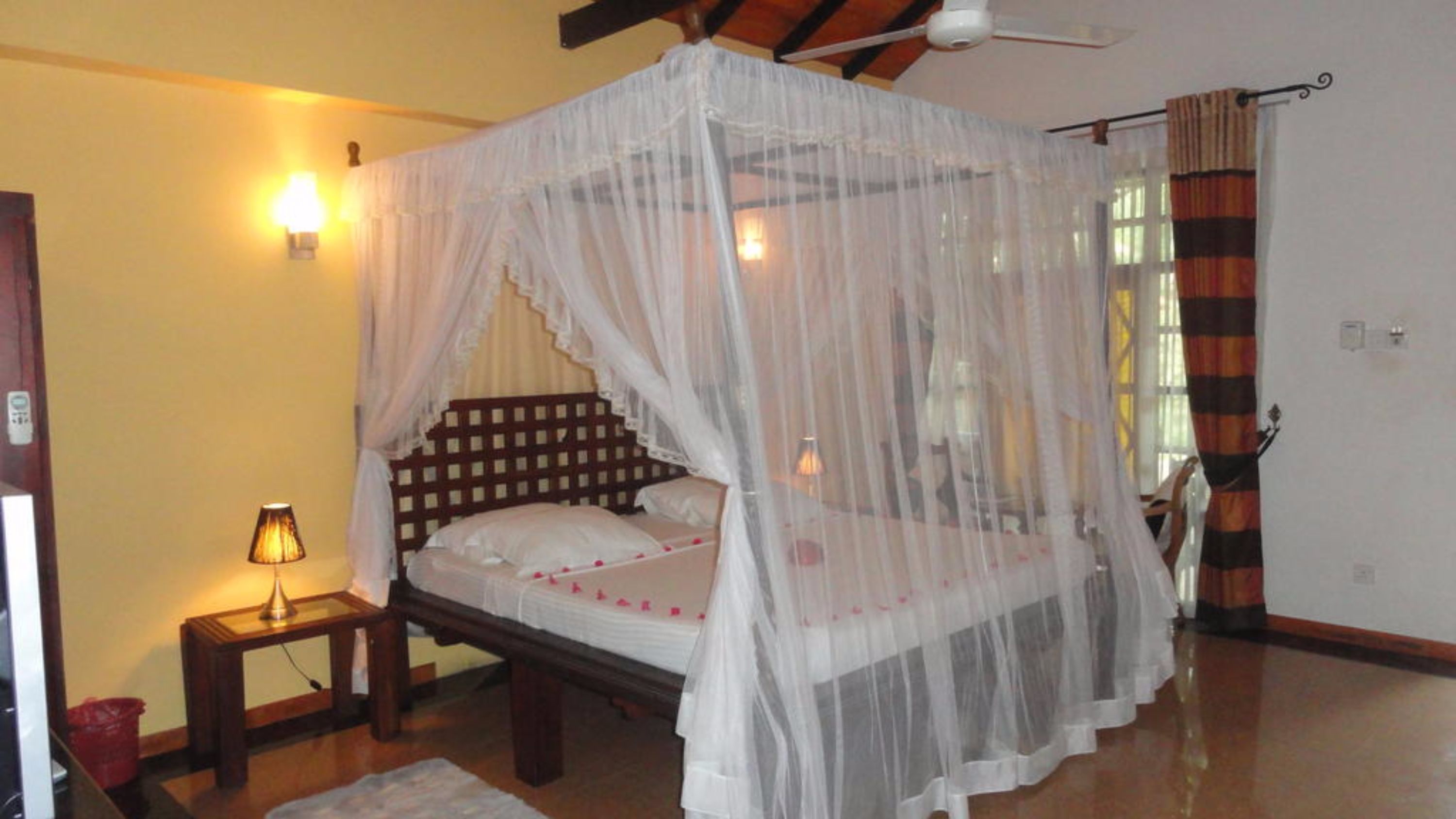 A bedroom with a four-poster canopy bed, a bedside table with a lamp, and a window with curtains.