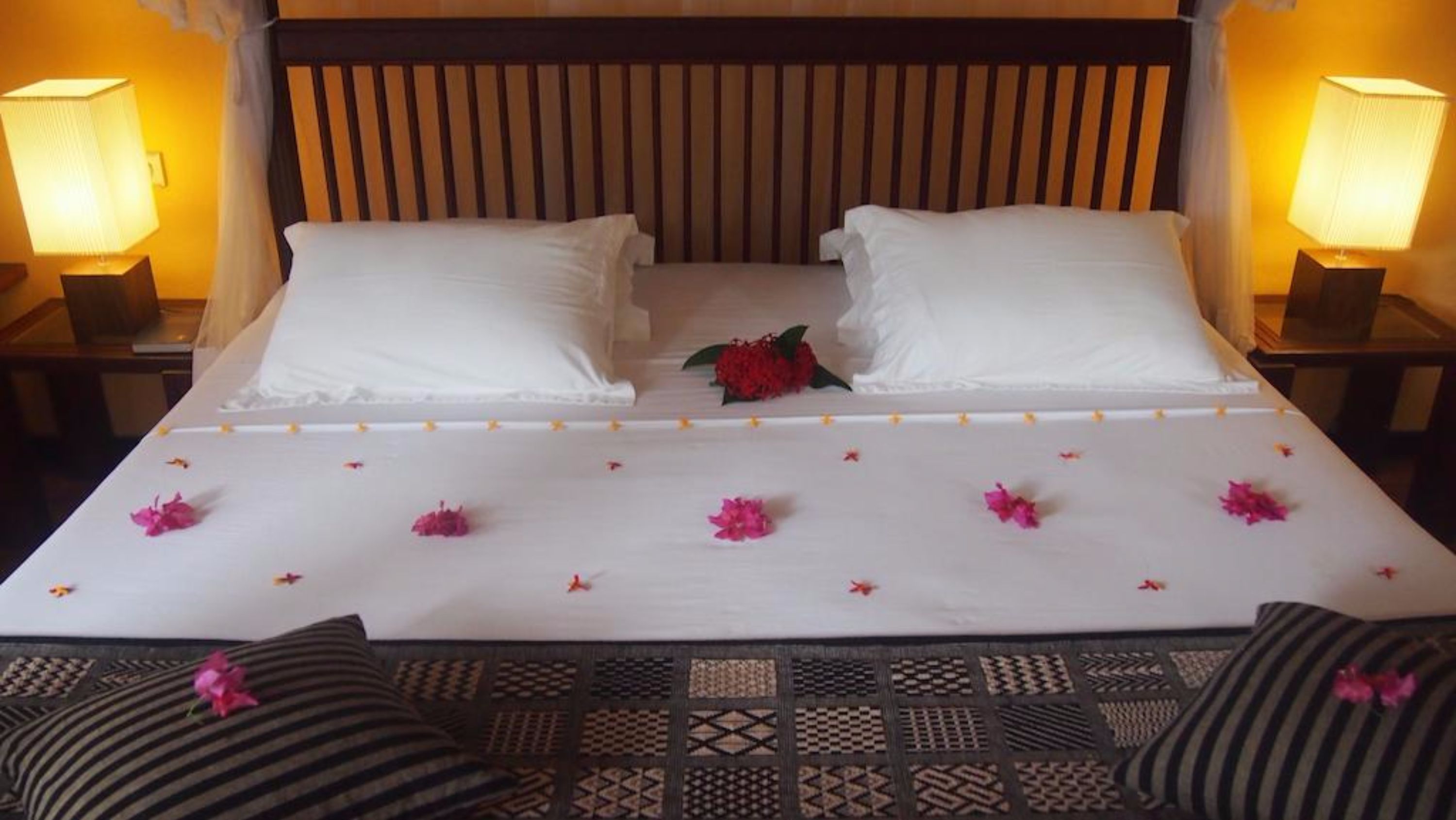 A neatly made bed with white linens, two pillows, and a decorative red flower centerpiece.