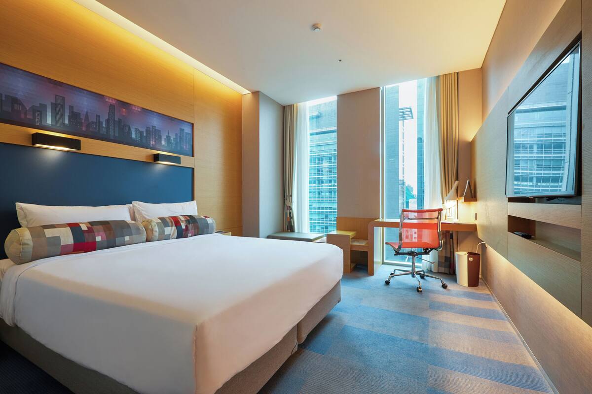 Ein Hotelzimmer mit einem großen Bett, einem Schreibtisch, einem Stuhl und einem Stadtbild an der Wand.