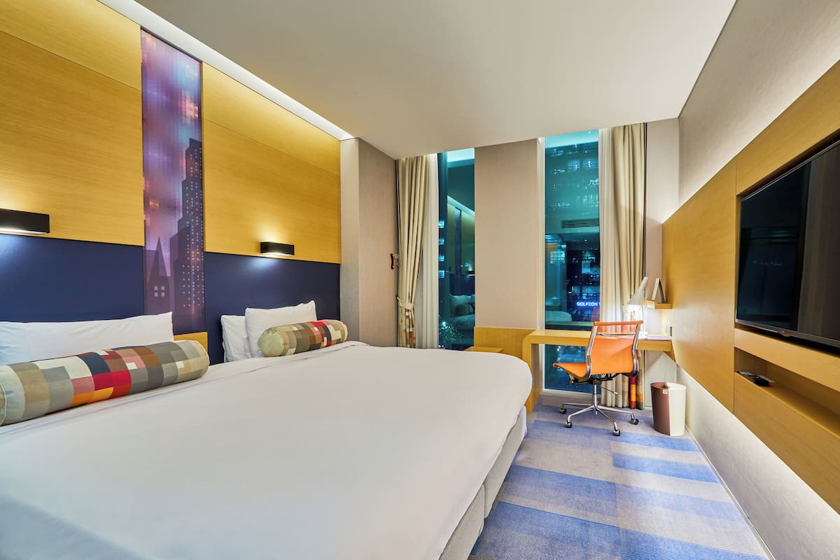 Ein Hotelzimmer mit einem großen Bett, einem Schreibtisch mit Stuhl, einem Fernseher und Blick auf eine Stadtansicht durch das Fenster.