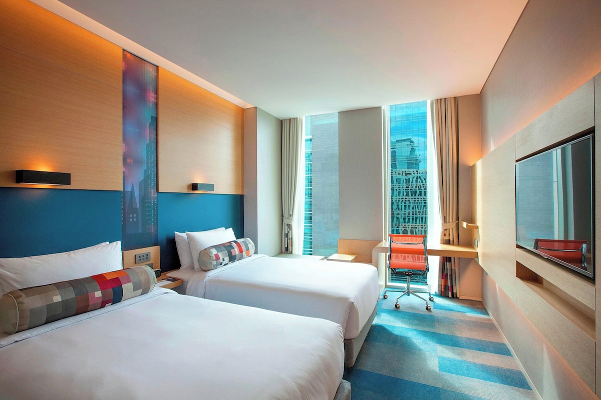 Ein Hotelzimmer mit zwei Betten, einem Schreibtisch, einem Sessel, einem Fernseher und einem großen Fenster mit Blick auf die Stadt.