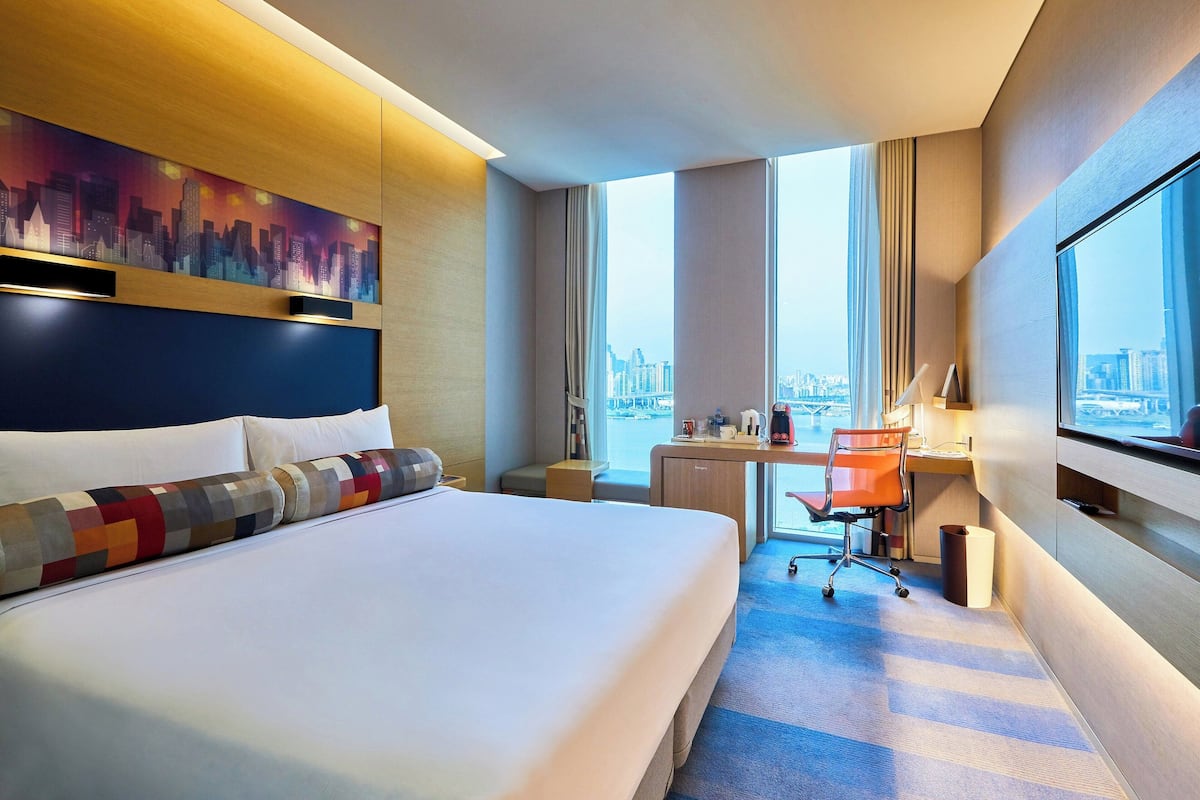 Ein Hotelzimmer mit einem großen Bett, einem Schreibtisch mit Stuhl, Blick auf die Stadt und einem Stadtbild an der Wand.