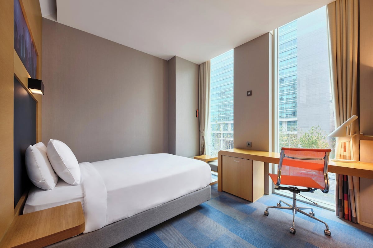 Ein Hotelzimmer mit Bett, Schreibtisch und Stuhl, großen Fenstern mit Blick auf die Stadt und einem blauen Teppich.