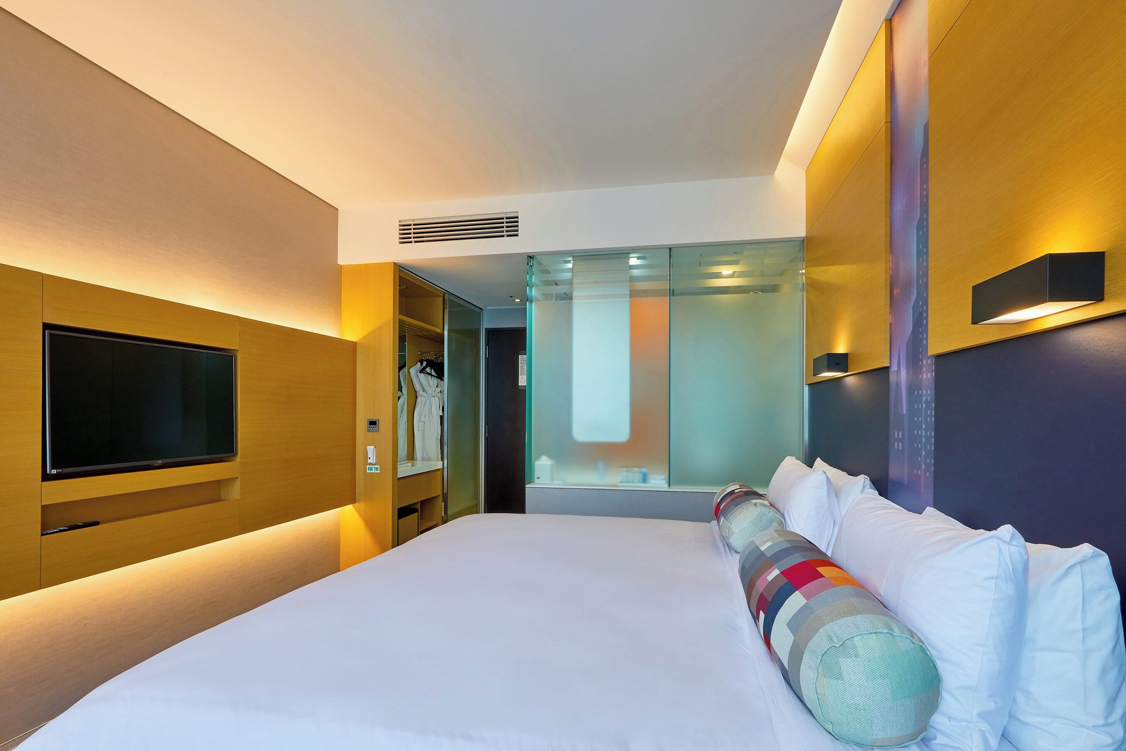 A modern hotel room with a large bed, a flat-screen TV, a wardrobe, and a glass partition.