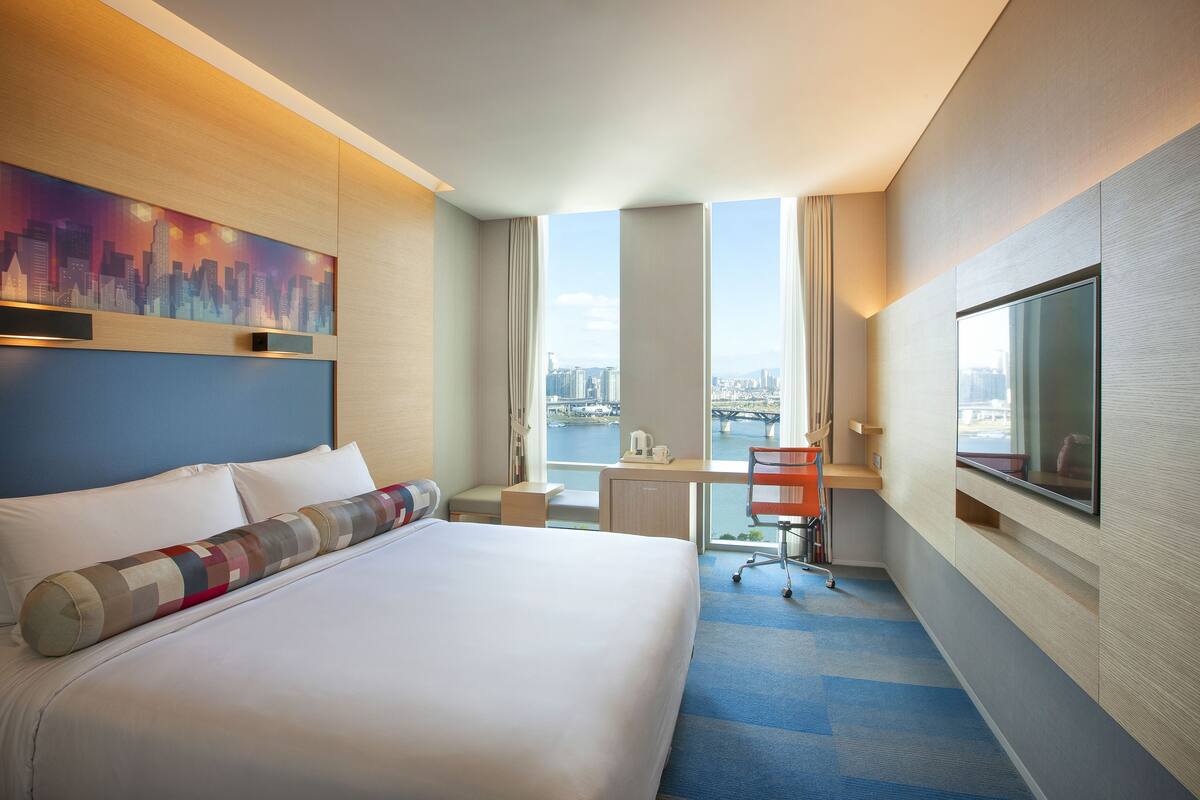 Ein Hotelzimmer mit einem großen Bett, einem Schreibtisch, einem Stuhl, einem Fernseher und Blick auf die Stadt.