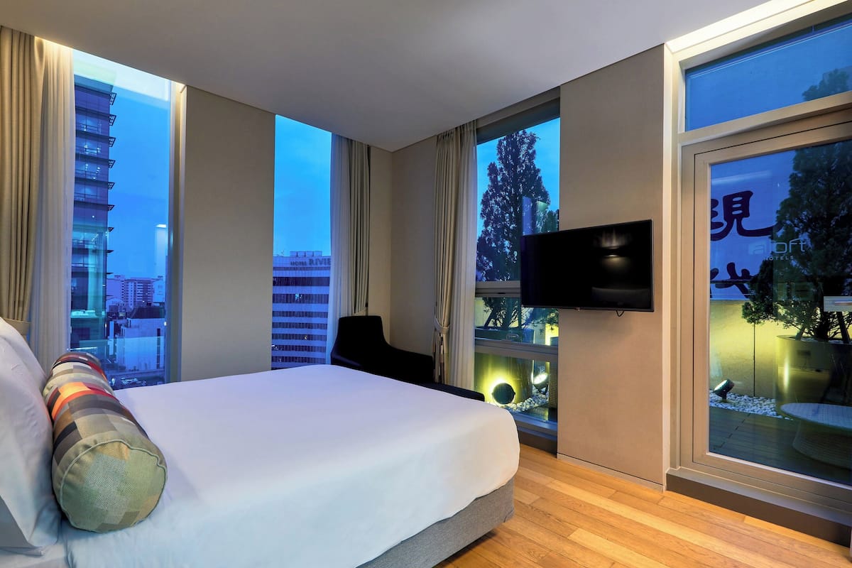 Ein Hotelzimmer mit einem großen Bett, einem an der Wand befestigten Fernseher und Blick auf die Stadt durch große Fenster.