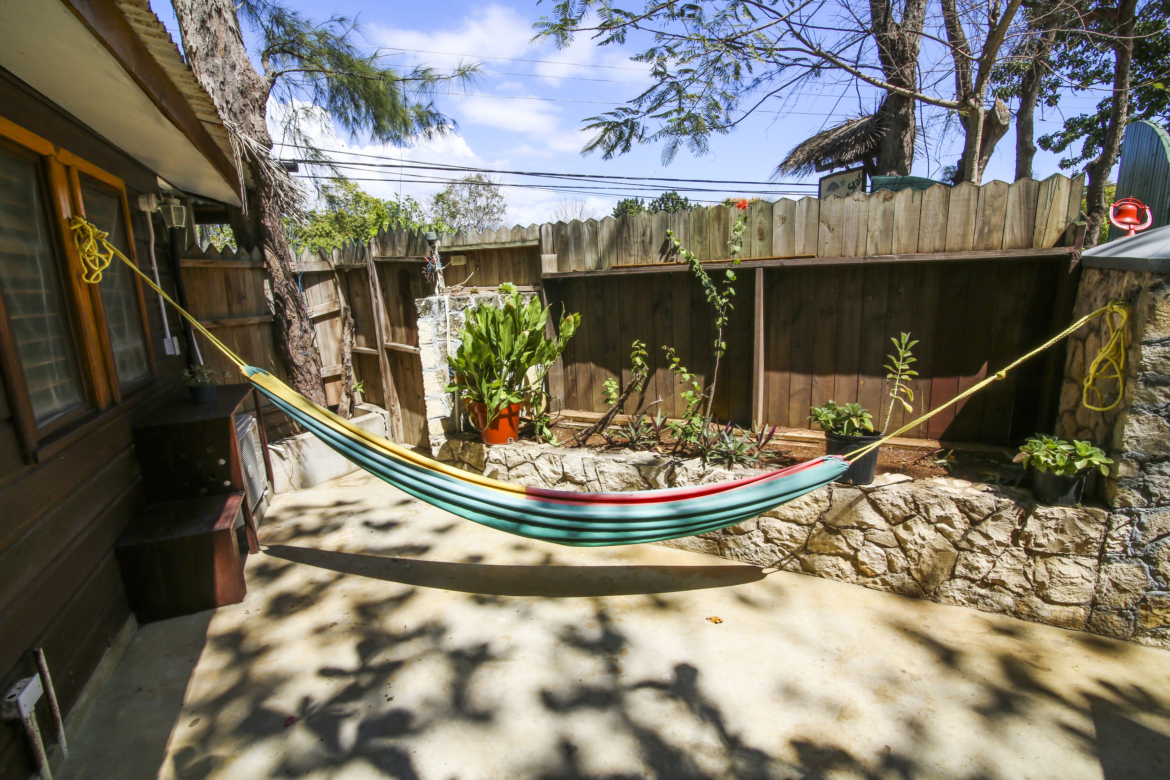 A hammock tied between two wooden posts in a backyard with a stone wall, potted plants, and a wooden fence.