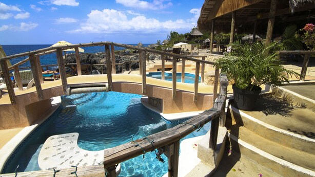 Zona de piscina con terraza de madera, una estructura con techo de paja y vista al océano.