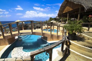 Ein Poolbereich mit Holzterrasse, einem strohgedeckten Bauwerk und Blick auf das Meer.